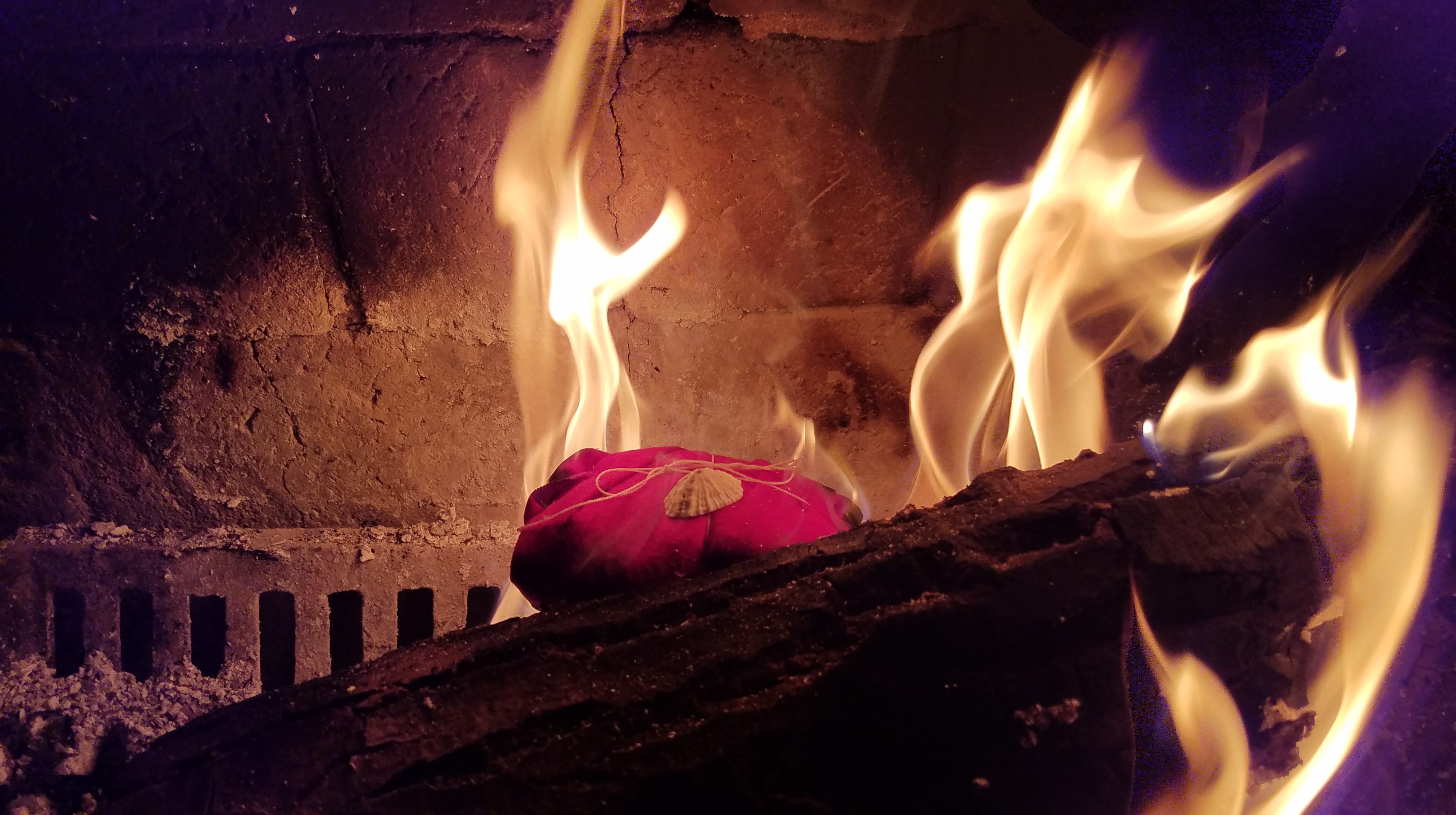 A pink cloth offering bundle tied with string and a shell, resting on a burning log in a fireplace