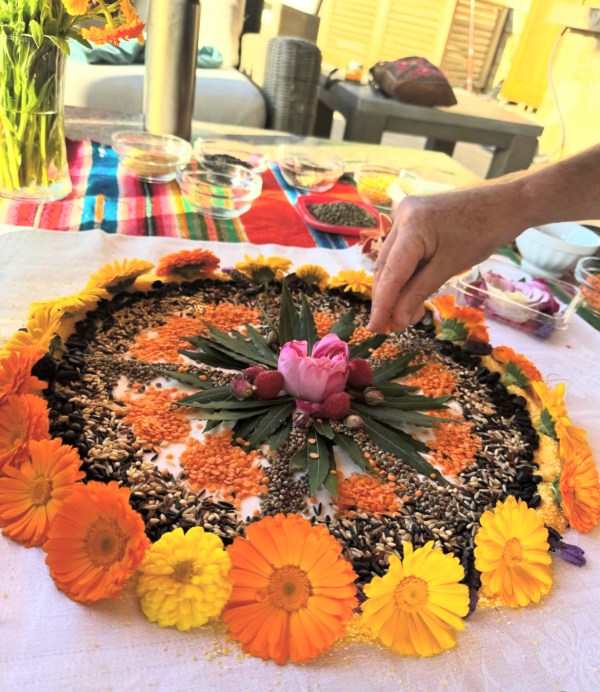 Hand placing seeds on a ceremonial mandala ringed with calendula, with a pink rose at the center