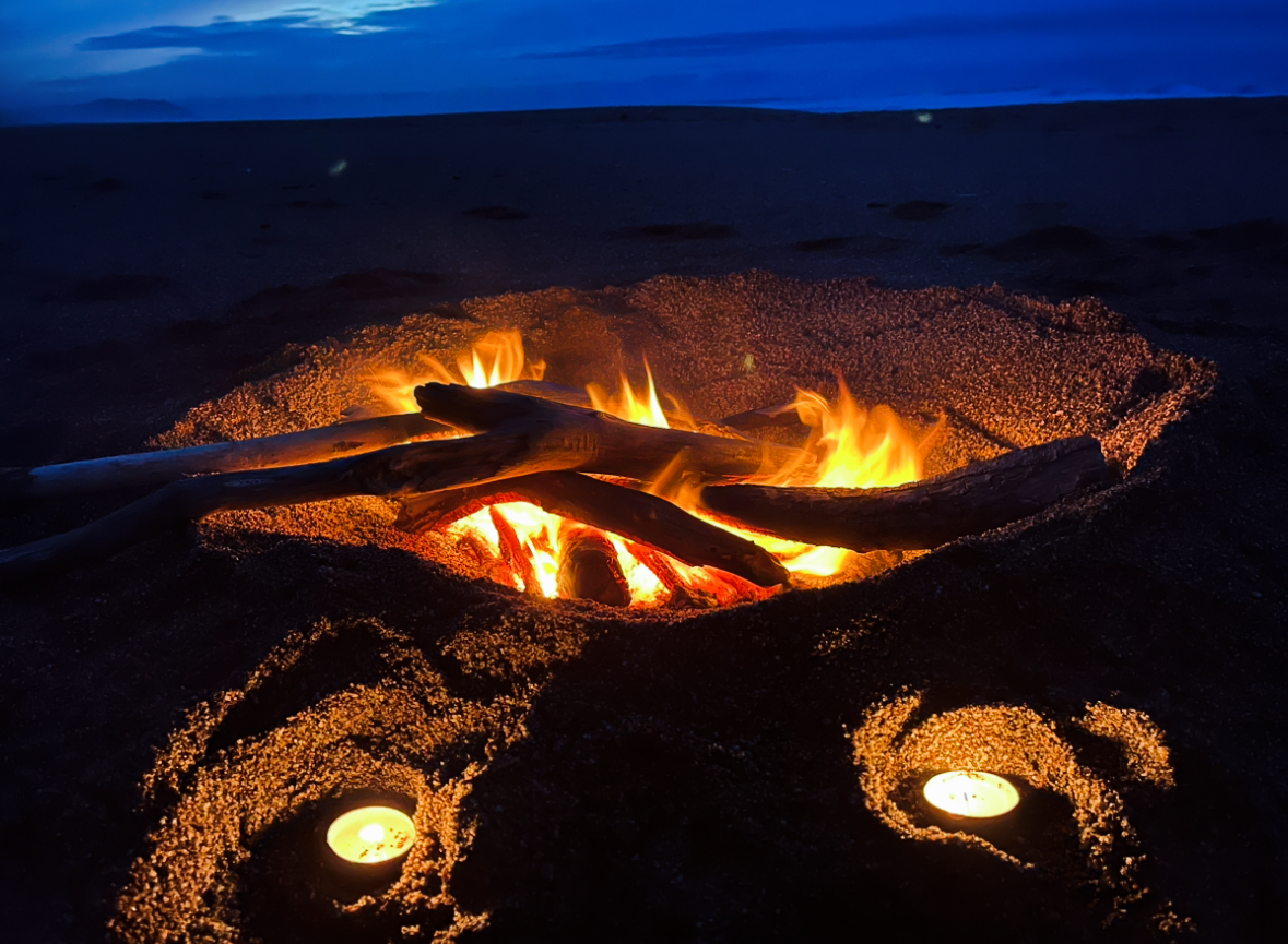 Driftwood fire burning in a sand pit on the beach at dusk, with two candles set in swirled sand in front